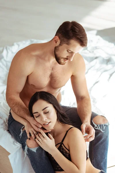Hombre guapo, musculoso y sin camisa abrazándose con hermosa mujer con los ojos cerrados en sujetador de encaje - foto de stock