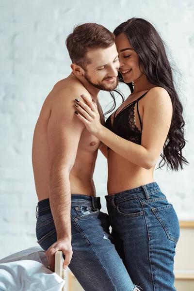 Hombre guapo y musculoso abrazándose con mujer hermosa y sonriente en sujetador de encaje en el dormitorio - foto de stock
