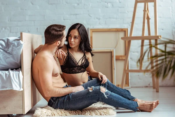 Handsome and muscular man sitting on rug and hugging with beautiful woman in lace bra in bedroom — Stock Photo