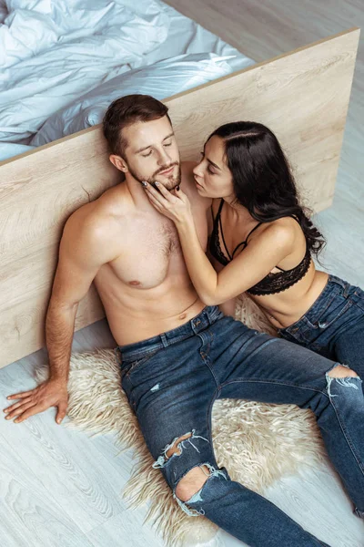 Hombre guapo y musculoso con los ojos cerrados sentado en la alfombra y abrazando con hermosa mujer en sujetador de encaje en el dormitorio - foto de stock