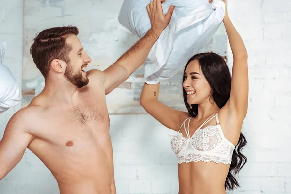 Muscoloso e bello uomo che tiene cuscini e gioca con bella donna in reggiseno di pizzo in camera da letto — Foto stock