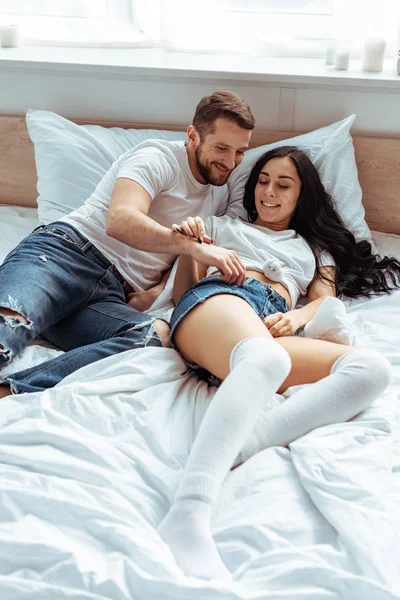 Hombre guapo en jeans y hermosa mujer en camiseta blanca acostado y sonriendo en el dormitorio - foto de stock