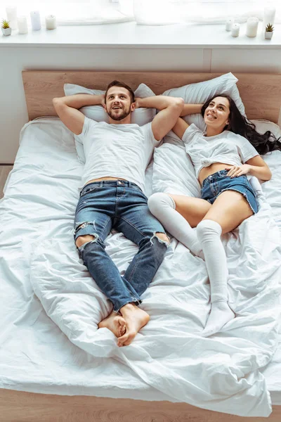 Hombre guapo en jeans y mujer hermosa y sonriente en camiseta blanca mirando hacia arriba en el dormitorio - foto de stock