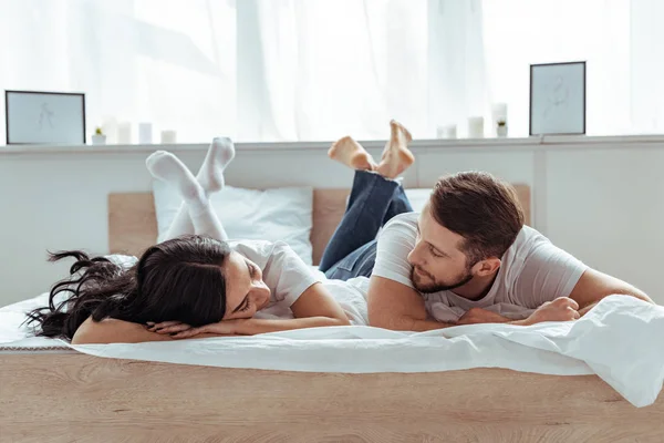 Парень в джинсах и женщина в шортах и футболке лежат на кровати и смотрят друг на друга в спальне — стоковое фото