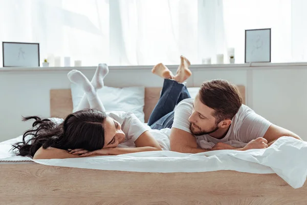 Freund in Jeans und Frau in Shorts und T-Shirt liegen auf dem Bett und schauen einander im Schlafzimmer an — Stockfoto