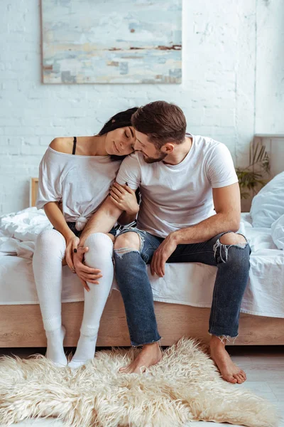 Hombre guapo en jeans y hermosa mujer sentada en la cama y abrazando - foto de stock