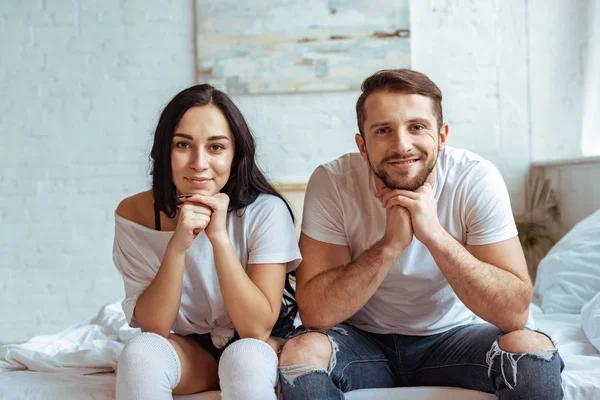 Handsome man in jeans and beautiful woman in t-shirt sitting on bed and looking at camera — Stock Photo
