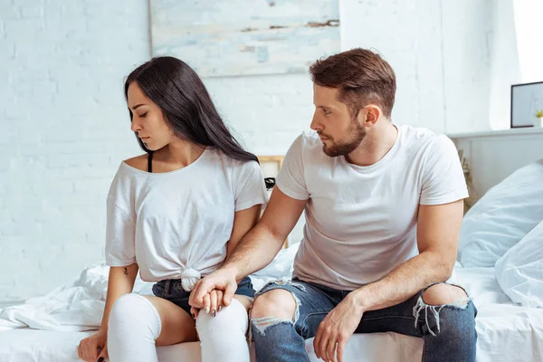 Handsome man in jeans holding hand of beautiful and sad woman in t-shirt sitting on bed — Stock Photo