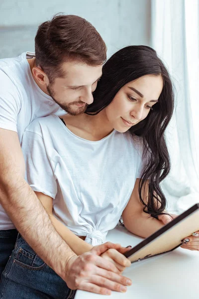 Guapo hombre abrazando hermosa y morena mujer en camiseta y sosteniendo foto - foto de stock