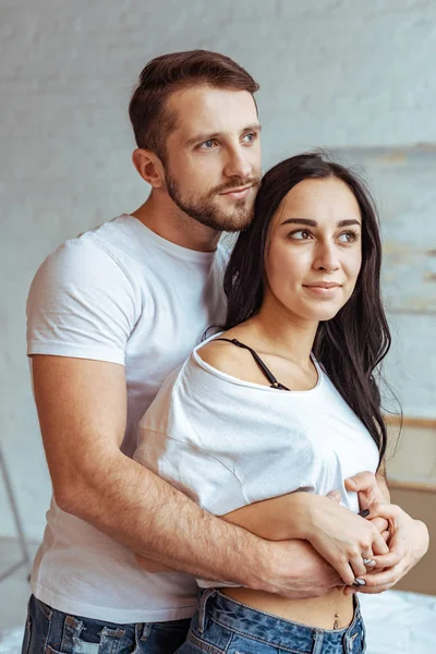 Hombre guapo abrazando hermosa y morena mujer en camiseta y mirando hacia otro lado - foto de stock