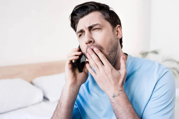 Hombre guapo bostezando mientras habla en el teléfono inteligente en el dormitorio - foto de stock