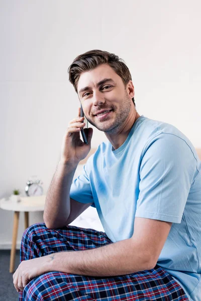 Hombre guapo en pijama hablando en teléfono inteligente y mirando a la cámara - foto de stock
