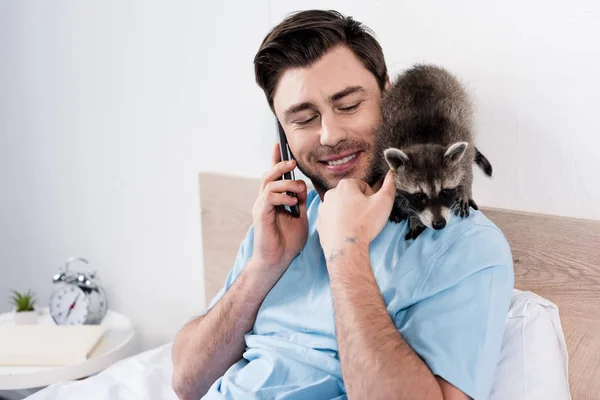 Sonriente hombre guapo hablando en el teléfono inteligente mientras mapache divertido sentado en su hombro - foto de stock