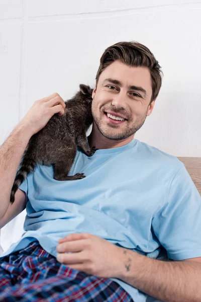 Fröhlich gutaussehender Mann kuschelt mit niedlichem Waschbär und schaut in die Kamera — Stockfoto