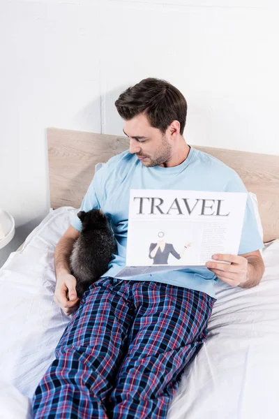 Schöner Mann kuschelt im Bett mit entzückendem Waschbär und hält Reisezeitung in der Hand — Stockfoto