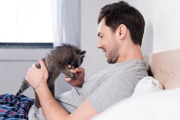 Selective focus of cheerful man playing with adorable raccoon in bedroom — Stock Photo