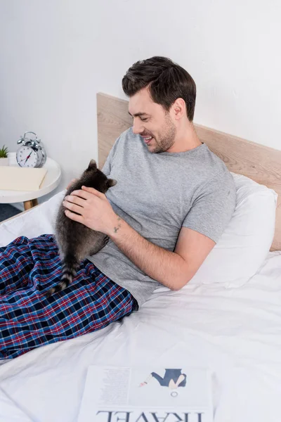 Bello uomo sorridente in pigiama rilassante a letto con procione divertente — Foto stock