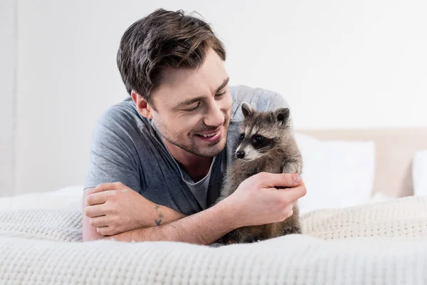 Handsome smiling man resting on bedding with adorable raccoon — Stock Photo