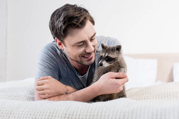 Bonito sorrindo homem descansando na cama com adorável guaxinim — Fotografia de Stock