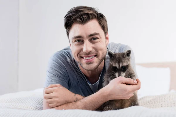 Cheerful man cuddling with adorable raccoon on bedding and looking at camera — Stock Photo