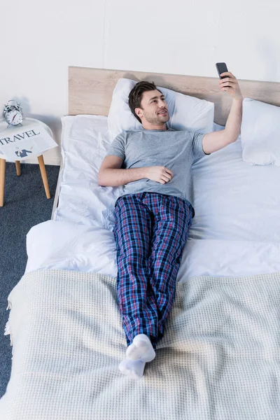Hombre guapo tomando selfie con teléfono inteligente mientras descansa en la cama en casa - foto de stock
