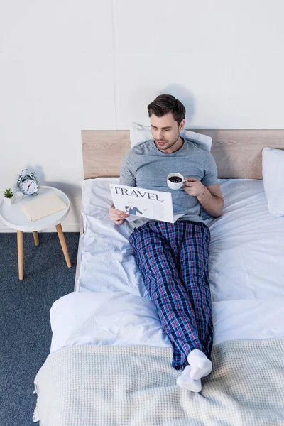 Красивий чоловік у піжамах відпочиває на ліжку з чашкою кави та туристичною газетою — стокове фото
