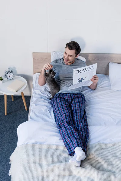 Веселый человек играет со смешным енотом, лежа на кровати с газетой о путешествиях — стоковое фото