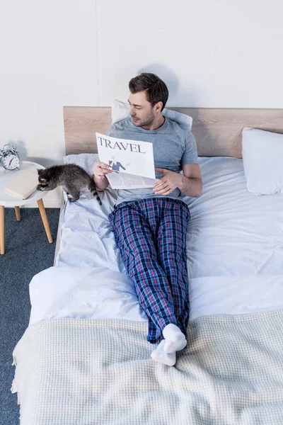Красивый мужчина лежит на кровати с газетой о путешествиях рядом с очаровательным енотом — стоковое фото