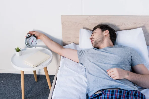 Schöner verschlafener Mann hält Wecker, während er zu Hause auf Bettwäsche liegt — Stockfoto