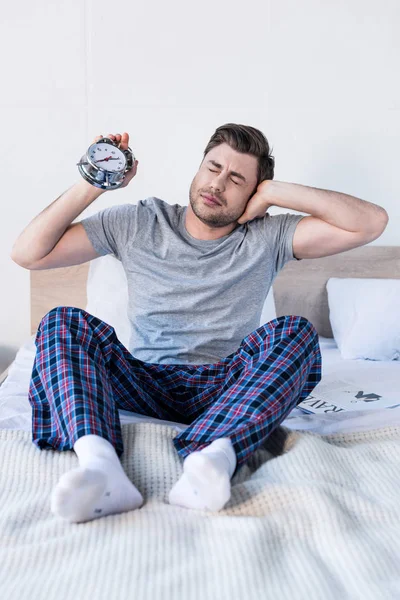 Schöner Mann dehnt und hält Wecker, während er auf Bettwäsche sitzt — Stockfoto