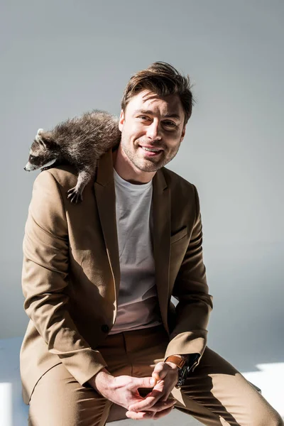 Happy man in beige suite, with funny raccoon on shoulder, smiling and looking at camera on grey — Stock Photo