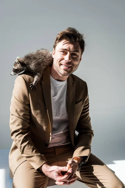 Homme heureux dans la suite beige, avec raton laveur drôle sur l'épaule, souriant et regardant la caméra sur gris — Photo de stock