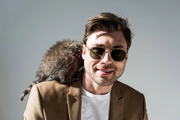 Smiling man in sunglasses with furry raccoon on shoulder on grey — Stock Photo