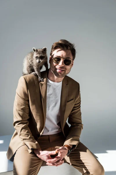 Handsome man in sunglasses with cute raccoon on shoulder on grey — Stock Photo