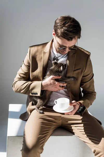 Красивий чоловік в сонцезахисних окулярах обнімається чарівним єнотом, тримаючи чашку кави — стокове фото