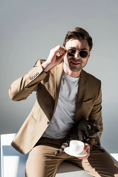 Красивый мужчина со смешным енотом и кофейной чашкой на коленях, смотрящий в камеру на сером — стоковое фото