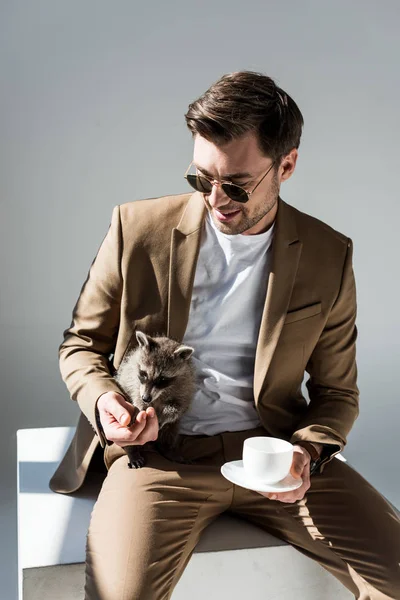 Cheerful handsome man sitting on sunlight and holding funny raccoon and coffee cup — Stock Photo