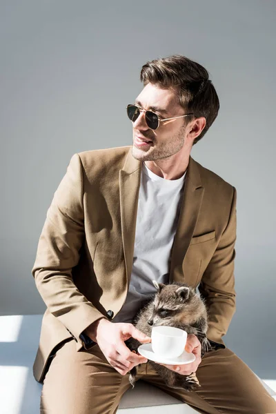 Smiling man sitting on sunlight and holding funny raccoon and coffee cup on grey — Stock Photo
