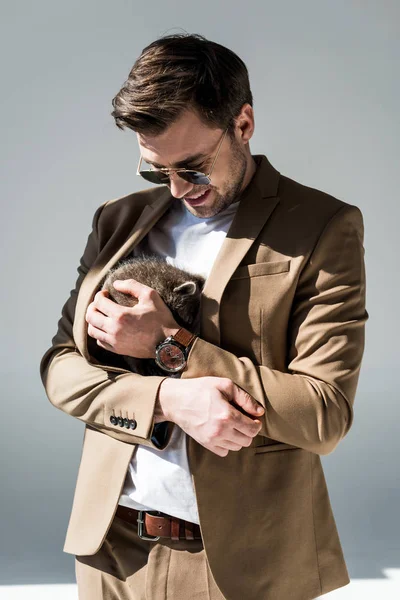 Handsome man in sunglasses cuddling with funny raccoon on grey — Stock Photo