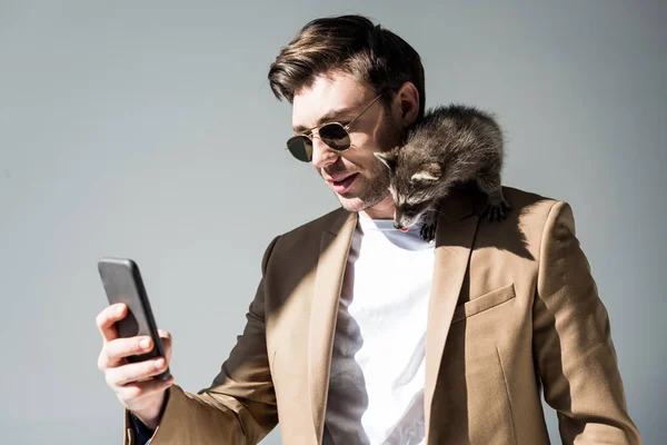 Гарний чоловік, з чарівним єнотом на плечі, приймаючи селфі зі смартфоном на сірому — стокове фото
