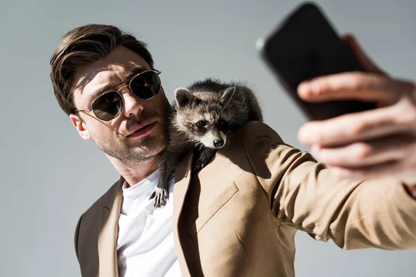 Enfoque selectivo del hombre guapo con mapache adorable en el hombro, tomar selfie con teléfono inteligente en gris - foto de stock