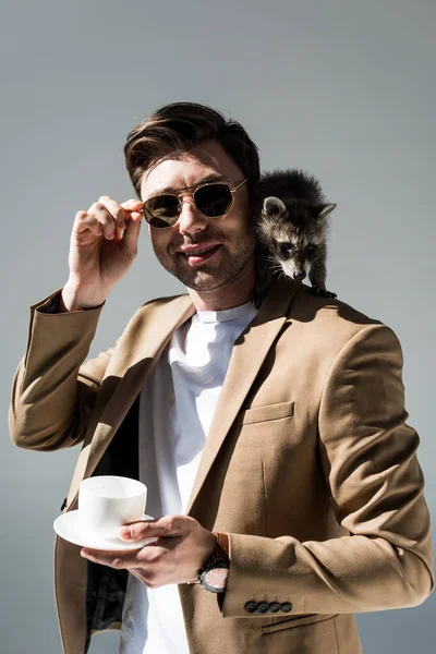 Sorrindo homem com guaxinim peludo no ombro, segurando xícara de café e olhando para a câmera no cinza — Fotografia de Stock