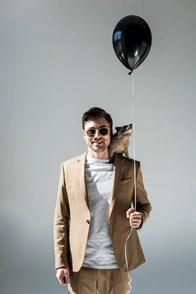 Sonriente hombre con divertido mapache en hombro sosteniendo globo de aire y mirando a la cámara en gris - foto de stock