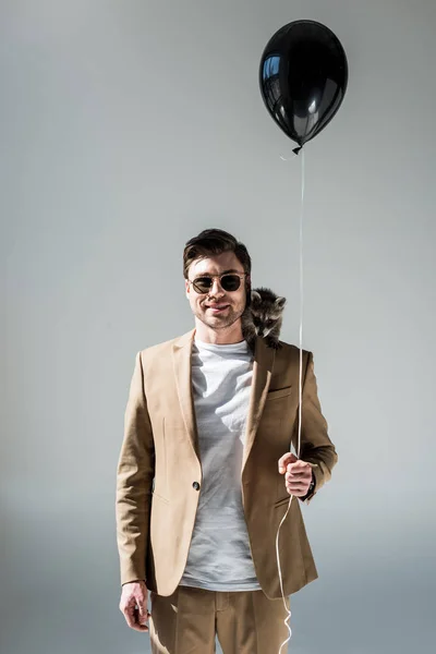 Schöner Mann mit niedlichen Waschbär auf der Schulter hält Luftballon und schaut in die Kamera auf grau — Stockfoto