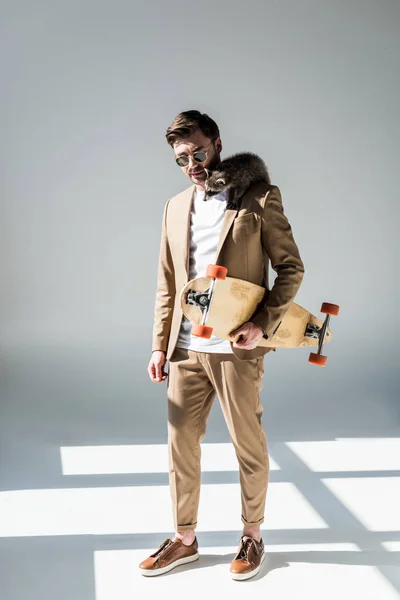 Handsome man with adorable raccoon on shoulder holding longboard on grey — Stock Photo