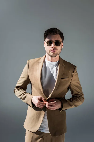 Handsome trendy man in beige suite and sunglasses looking at camera on grey — Stock Photo