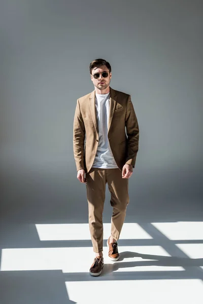 Hombre de buen aspecto elegante en suite beige y gafas de sol de pie en la luz del sol en gris - foto de stock