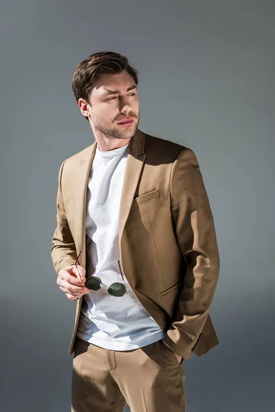 Handsome man in beige suite holding sunglasses and looking away on grey — Stock Photo