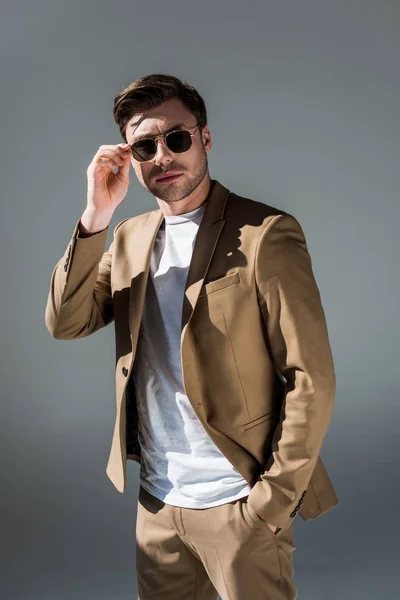 Bel homme à la mode en suite beige et lunettes de soleil regardant la caméra sur gris — Photo de stock