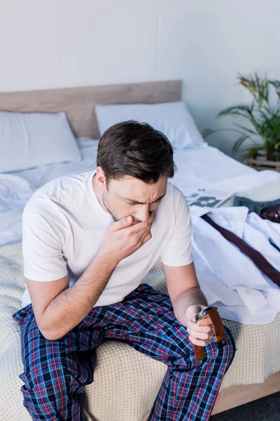 Bostezando hombre sosteniendo reloj mientras está sentado en la cama en casa - foto de stock